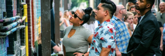 Multi-racial crowd look at something on a wall outside on a street.